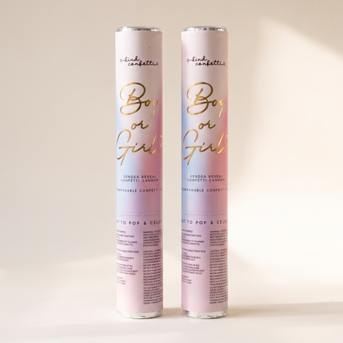 Boy or Girl gender reveal confetti cannon by Kind Confetti, two pastel pink and blue tubes with gold script lettering standing upright on a cream background in soft sunlight.