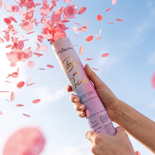 Gender reveal confetti cannon being popped against a blue sky, two hands holding the tube as a burst of pink biodegradable tissue paper confetti sprays into the air.