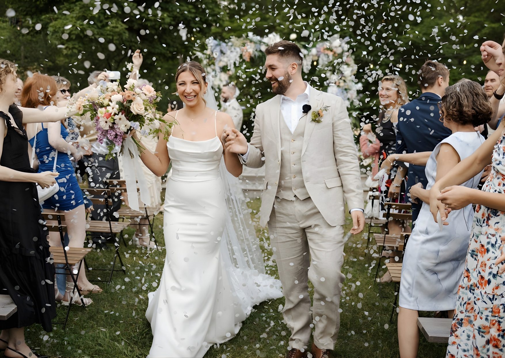 Wedding couple walking through confetti with guests on a grassy area.