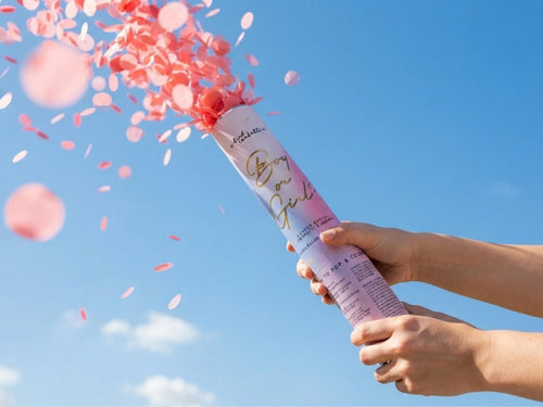 Hand firing a gender reveal cannon, pink biodegradable rice paper confetti bursting against a blue sky