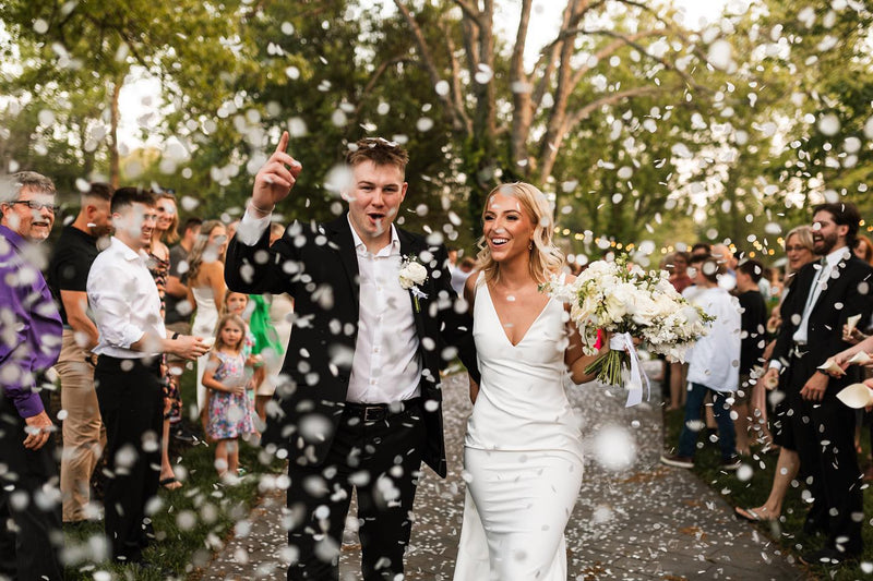 Wedding couple walking through confetti with guests on a path outdoors.
